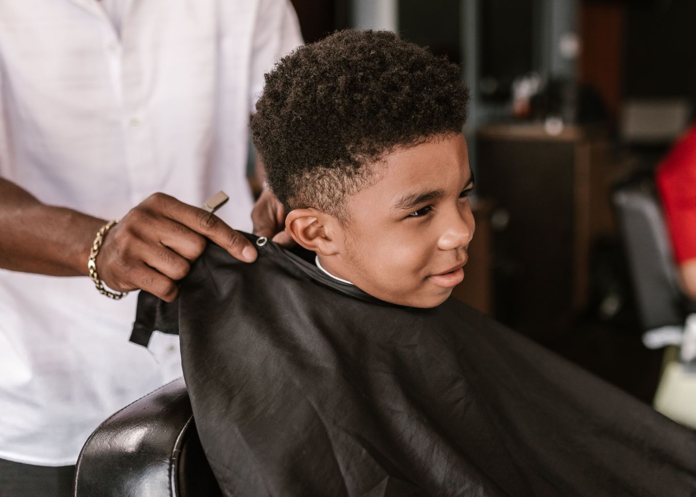 A young boy happily getting a haircut inside a modern barbershop, observed by an adult.